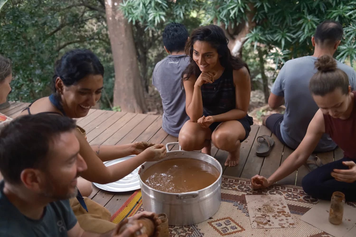 Residents sitting on a shaded wooden deck around a large pot, cups in hand