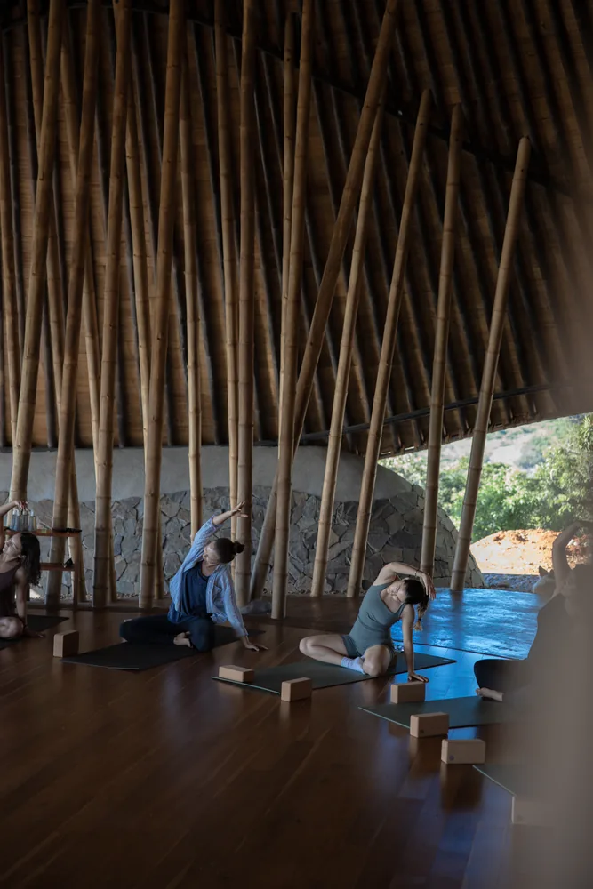 Two people stretching on mats under the bamboo maloca roof, hills beyond