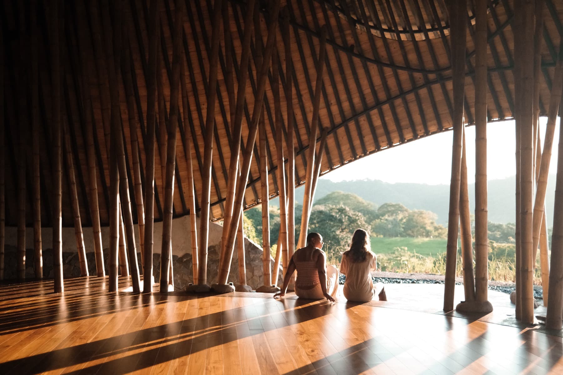 Two residents sitting on the maloca floor at golden hour, looking out at the hills